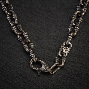 May include: A silver necklace with an intricate chain design. The necklace has an ornate clasp and a circular pendant with a symbol. The chain links have a textured appearance, and the piece is set against a dark background.