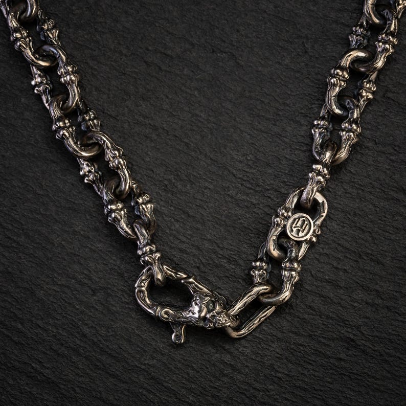 May include: A silver necklace with an intricate chain design. The necklace has an ornate clasp and a circular pendant with a symbol. The chain links have a textured appearance, and the piece is set against a dark background.