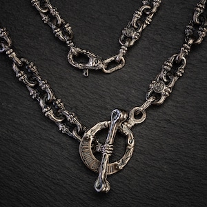 May include: A silver-coloured necklace with a distinctive toggle clasp shaped like a bone. The chain links have an ornate, textured design. The clasp has the text "US MORTIS" engraved on it. The necklace is displayed on a dark, textured surface.