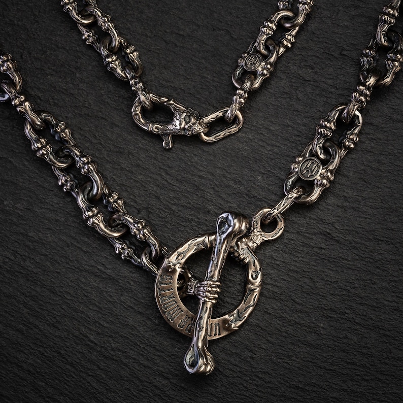 May include: A silver-coloured necklace with a distinctive toggle clasp shaped like a bone. The chain links have an ornate, textured design. The clasp has the text "US MORTIS" engraved on it. The necklace is displayed on a dark, textured surface.