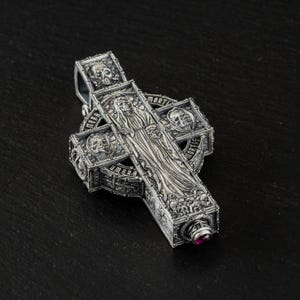 May include: Detailed silver pendant in the shape of a cross. The cross features intricate carvings of skulls, a robed figure, and the words "Umbra Mortis". A small, round, red gemstone is set at the bottom.