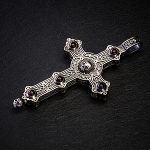 May include: A silver cross pendant with intricate detailing and five red gemstone accents. The center of the cross features a skull design.