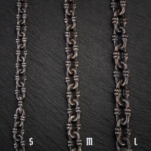 May include: Three silver-coloured metal chains of varying lengths, labelled S, M, and L, are displayed against a dark, textured background. The chains have a detailed, ornate design with interconnected links. The chains are likely jewellery, such as necklaces or bracelets.