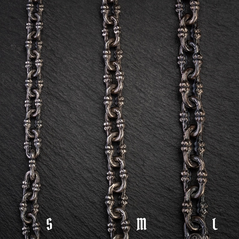 May include: Three silver-coloured metal chains of varying lengths, labelled S, M, and L, are displayed against a dark, textured background. The chains have a detailed, ornate design with interconnected links. The chains are likely jewellery, such as necklaces or bracelets.