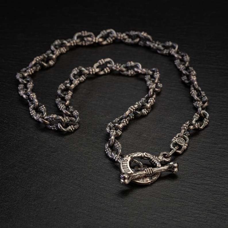 May include: A silver chain necklace with a toggle clasp. The chain links have an intricate design. The clasp features the word "MORTIS" and a bone-shaped bar. The necklace is shown on a dark surface.
