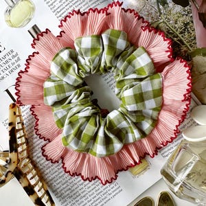 May include: A green and white gingham hair scrunchie with a pink ruffled trim. The scrunchie is on a book with text about a berry.