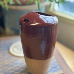 May include: A handmade ceramic vase with a two-tone design. The top portion is a deep reddish-brown, transitioning to a lighter tan color at the base. The vase has a cylindrical shape with a small hole near the top. The vase is approximately 6 inches tall.