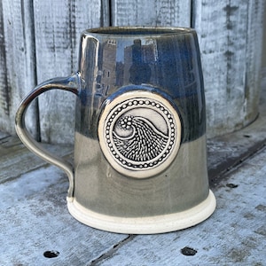 May include: A ceramic mug with a blue and gray glaze. The mug has a white rim and a white circle with a wave design in the center.