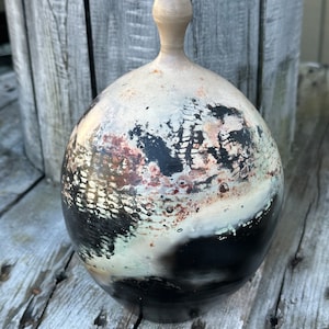 May include: A round, ceramic vase with a black, white, and brown speckled glaze. The vase has a narrow neck and a wide, rounded body.