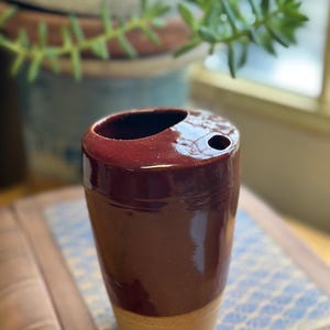 May include: A handmade, cylindrical ceramic toothbrush holder. The top is a deep burgundy color with two holes, and the bottom is a natural beige. The holder is on a patterned surface, with greenery in the background.