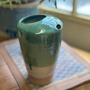 May include: A handmade ceramic tumbler with a unique design. The top is a deep teal color, transitioning to a lighter green and beige base. It features a small hole near the rim, suggesting a straw or utensil holder. The tumbler sits on a patterned surface.
