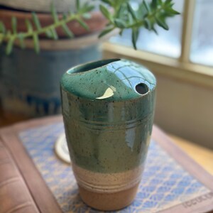 May include: A handmade ceramic vase with a unique design. The vase is cylindrical, with a teal green upper section and a beige lower section. It has a small opening on top and a textured surface. The vase is sitting on a book with a blue and white pattern.