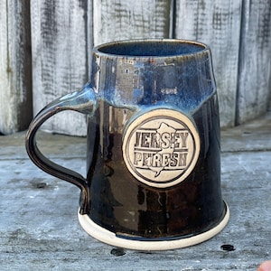 May include: A dark blue ceramic mug with a white rim and a white circle logo that says "JERSEY FRESH" with a stylized outline of the state of New Jersey.