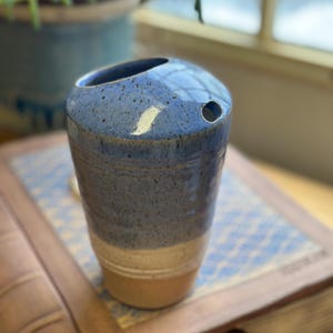 May include: A handmade ceramic vase with a blue speckled glaze. The vase has a cylindrical shape with a rounded top and a small hole on the side. The bottom is a light tan color.