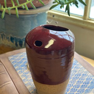 May include: A handcrafted, cylindrical vase with a deep burgundy glaze on the upper portion and a speckled tan base. The vase has a circular opening and a small hole on the side. The pottery is likely ceramic.