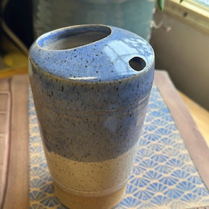 May include: A handmade ceramic tumbler with a speckled blue and cream glaze. The cylindrical cup has a small hole near the top and is sitting on a patterned coaster. The pottery is likely for home decor.