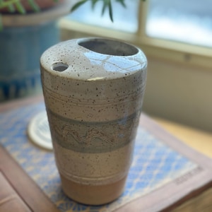 May include: A speckled ceramic tumbler with a rounded top and a small hole. The tumbler features a light beige base, a band of blue-grey waves, and a speckled off-white top. The design is simple and elegant, suitable for a variety of uses.