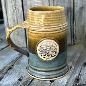 Puede incluir: Una jarra de cerveza de cerámica marrón y azul con un diseño circular blanco que presenta una ilustración en blanco y negro de un tren. La jarra tiene un asa y una base redondeada.