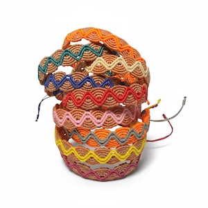May include: A stack of woven bracelets featuring a tan base with a repeating scalloped pattern. Each bracelet is accented with a distinct wavy thread in colors including orange, teal, cream, blue, red, pink, gray, yellow, and magenta.