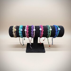 May include: A collection of seven macrame bracelets arranged on a black display stand. Each bracelet features a woven leaf-like pattern in various colors including beige, turquoise, pink, purple, green, and blue, accented with a single round bead.