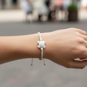 May include: A white turtle charm bracelet. The bracelet features a small, detailed turtle pendant and white beads strung on a beige cord. The bracelet is adjustable with sliding knots.