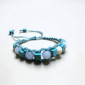 May include: A handmade macrame bracelet featuring a light blue woven band. The bracelet is adorned with square turquoise beads, round light blue beads, and a single white bead. The adjustable closure is also light blue.