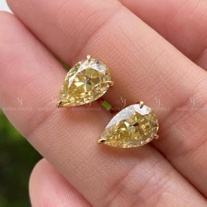May include: A pair of pear-cut yellow gemstones set in gold metal stud earrings. Each stone features a faceted surface that reflects light, showcasing a vibrant golden-yellow hue. The earrings are secured with gold posts and backings.