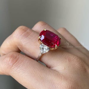 May include: A ring featuring a large, oval-cut vibrant red gemstone set in yellow gold prongs, accented by a single, smaller, triangular-cut clear white diamond on the side, mounted on a polished, slender, metallic band.