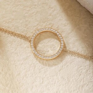 May include: A gold-toned chain bracelet featuring a central circular pendant. The circular pendant is encrusted with a series of small, clear, sparkling stones along its entire outer edge. The chain is delicate and connects to both sides of the circle.