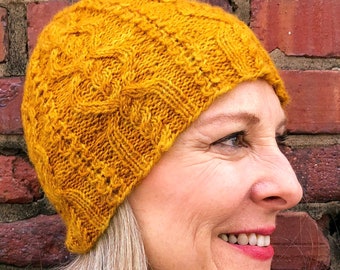 May include: A woman wearing a mustard yellow knitted hat with a cable knit pattern. The hat is pulled down over her forehead and she is smiling.