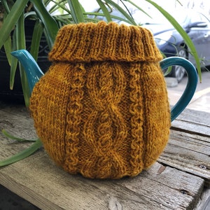May include: A knitted teapot cozy in a mustard yellow color with a cable knit pattern. The cozy is on a teal teapot.