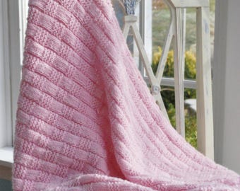 May include: A pink knitted baby blanket with a cable knit pattern, draped over a white chair.