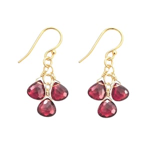 May include: A pair of gold dangle earrings, each featuring three faceted, teardrop-shaped deep red garnet gemstones suspended from a gold wire hook. The stones are individually wire-wrapped and attached to a central gold ring, creating a cluster.