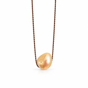 May include: A necklace featuring a single, large, irregular-shaped, iridescent peach-colored pearl suspended on a thin, dark brown twisted cord. The pearl has a smooth, lustrous surface with subtle variations in tone, hanging centrally from the cord.