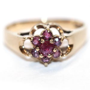 Può includere: Un anello in oro con un gruppo centrale di sette pietre preziose rotonde di colore rosso rosato intenso disposte a fiore. Le pietre sono incastonate in una montatura in oro con dettagli traforati sui lati della fascia dell'anello.