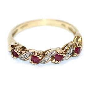 May include: A gold ring featuring a repeating pattern of red gemstones and small clear stones. The ring has a twisted design, with the gemstones set in the center of each twist. The ring is set against a white backdrop.