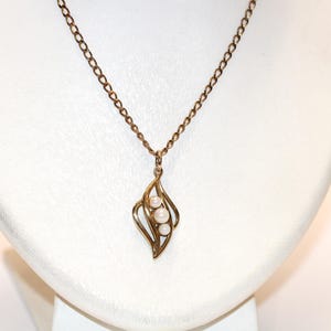 Könnte beinhalten: Eine goldfarbene Halskette mit einer feinen Kette und einem Anhänger in geschwungenem, durchbrochenem Design. Der Anhänger ist mit drei kleinen, runden, weißen Perlen verziert, die in einer vertikalen Linie in den Metallkurven angeordnet sind.