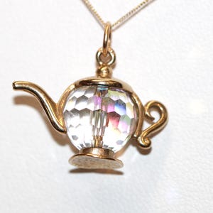 May include: A gold-toned metal pendant shaped like a teapot, featuring a faceted clear crystal body that reflects light with iridescent hues. The pendant includes a curved spout, a small handle, and a circular base, suspended from a thin metal chain.