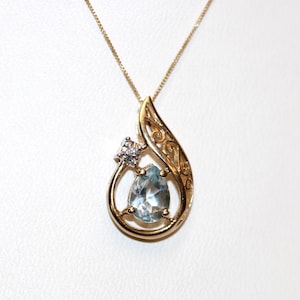 May include: A gold pendant necklace featuring a pear-shaped light blue gemstone set within a swirling gold frame. A small, square-cut clear stone is positioned above the blue gem. The pendant hangs from a delicate gold chain with a fine link design.