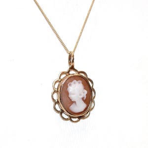 May include: A gold pendant necklace featuring an oval-shaped cameo with a white profile carving of a person against a light brown background. The cameo is set within a scalloped gold frame and hangs from a thin, delicate gold chain.