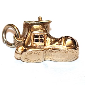 May include: A gold-toned metal charm shaped like a small house built into a shoe. The charm features a detailed roof, a square window with four panes, and a textured sole with circular patterns. A circular jump ring is attached to the back.