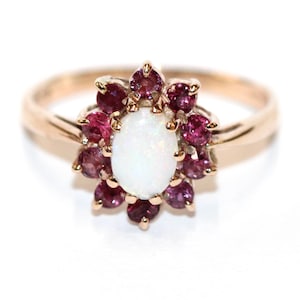 May include: A gold ring featuring a central oval white gemstone, surrounded by a cluster of smaller, round, pink gemstones. The ring's band is a simple, polished gold. The design is floral-inspired, with the pink stones resembling petals.