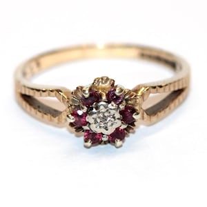 May include: A gold ring with a floral design. The centrepiece features a cluster of small, round, red gemstones around a central, smaller, white gemstone. The band has a textured pattern and a split design on each side of the floral centrepiece.