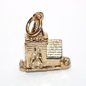 May include: A gold charm shaped like a small church building with a tall steeple, a textured brick exterior, and a tiled roof. The charm features a circular jump ring attached to the top of the steeple for hanging on a chain or bracelet.