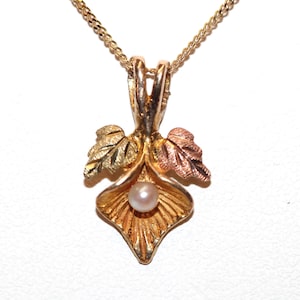 May include: A tri-colour gold pendant featuring a central white pearl set within a textured, fan-shaped leaf design. The pendant includes two smaller leaves in yellow and rose gold tones, suspended from a delicate gold chain necklace.