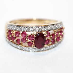 May include: A gold ring featuring a central oval-cut red ruby surrounded by smaller round-cut red rubies. The band is accented with two rows of small white diamonds along the top and bottom edges, creating a multi-stone layered design.