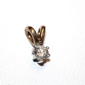 Pode incluir: Um pingente de metal prateado com uma pedra preciosa transparente de corte redondo fixada por quatro garras. O pingente inclui uma argola cónica e lisa no topo para fixação a uma corrente, apresentando um acabamento metálico polido.