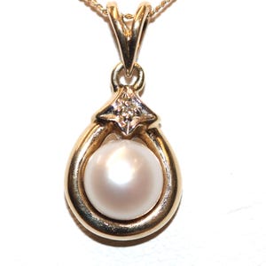May include: A gold pendant featuring a single round white pearl set within a teardrop-shaped gold frame. A small, clear, round-cut gemstone is mounted in a four-prong setting at the top of the pearl, connecting to a gold bail and chain.