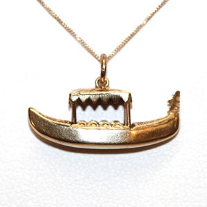 May include: A gold-toned metal pendant shaped like a gondola boat, featuring a small canopy with a scalloped edge and a pointed prow. The pendant is suspended from a delicate gold-toned chain that attaches to a small jump ring at the top centre.