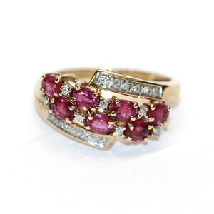 Può includere: Un anello d'oro con un gruppo di pietre preziose rosa ovali e piccoli diamanti trasparenti. L'anello ha un design curvo con le pietre preziose disposte in fila.